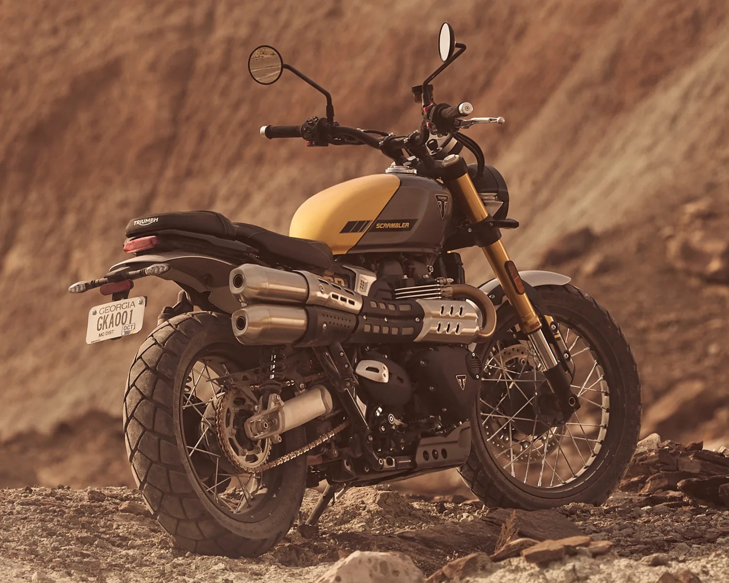 Yellow and gray Triumph Scrambler motorcycle parked on rocky terrain with dual exhaust pipes.