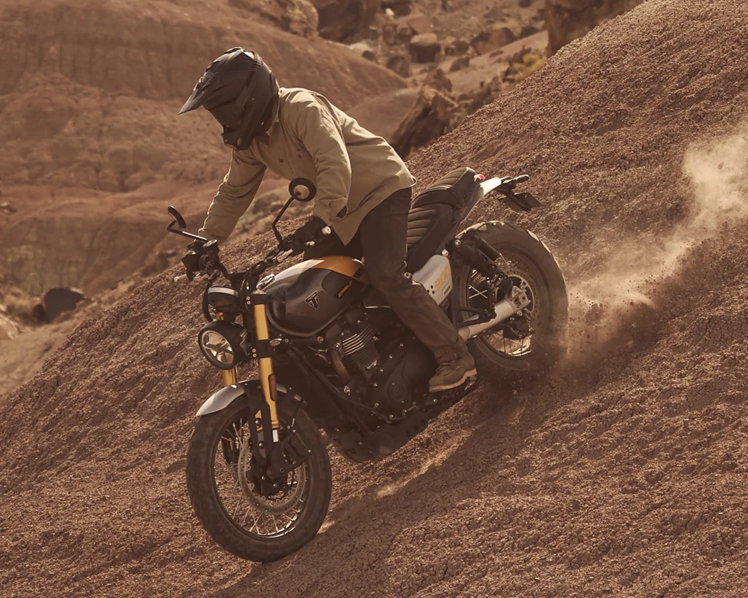Motorcyclist wearing a black helmet and beige jacket riding a black and yellow Triumph motorcycle on a dirt hill.