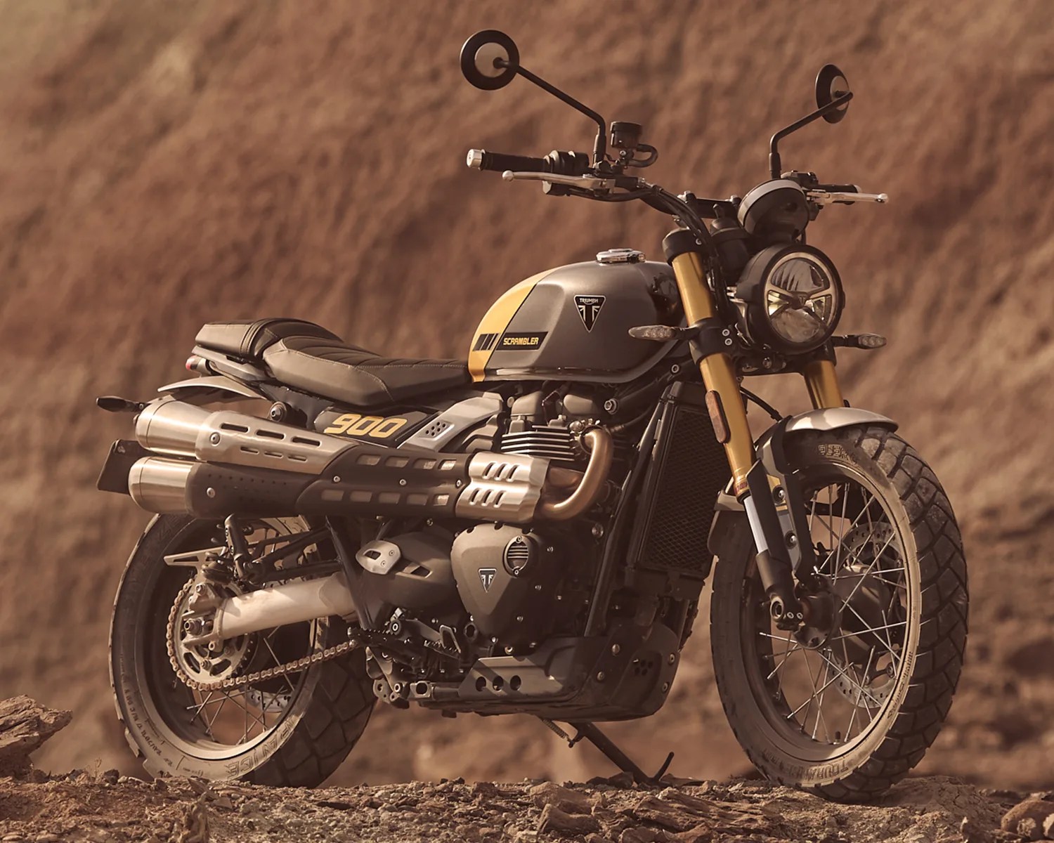 Matte gray and yellow Triumph Scrambler 900 motorcycle with dual exhaust pipes on a rocky terrain.