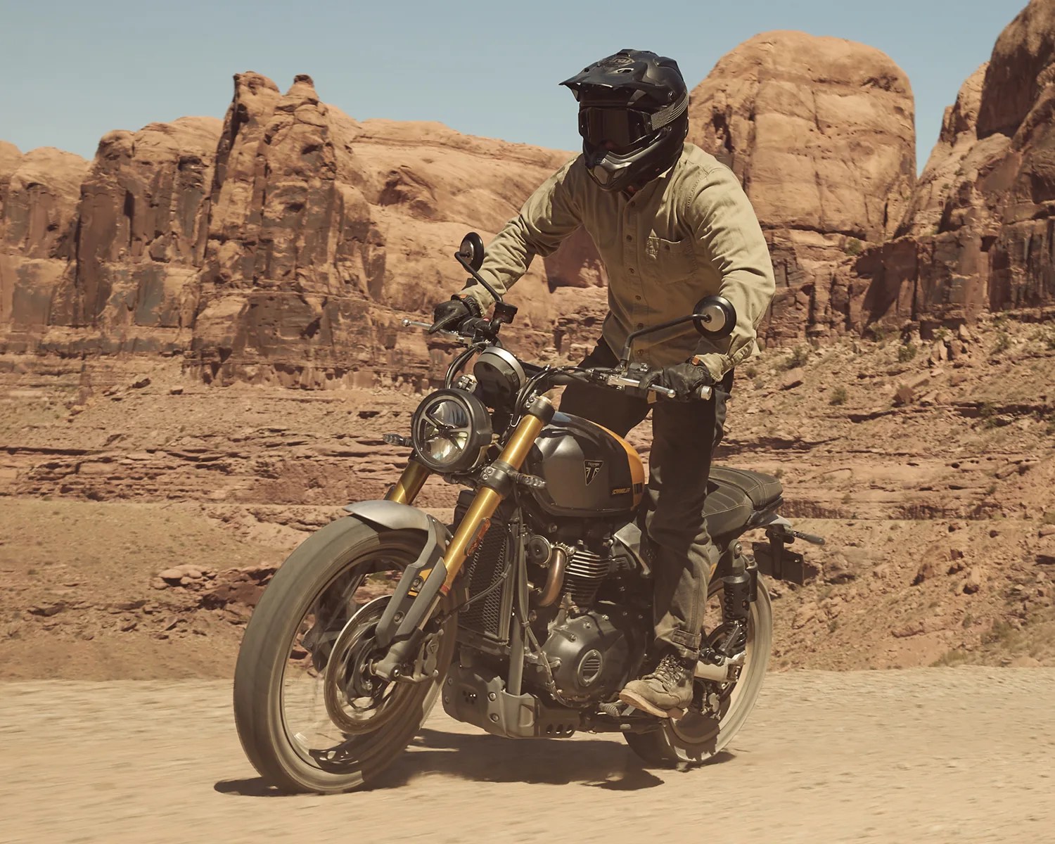 Person wearing a black helmet and tan jacket riding a gray and orange Triumph motorcycle on a dirt road with rocky cliffs in the background.