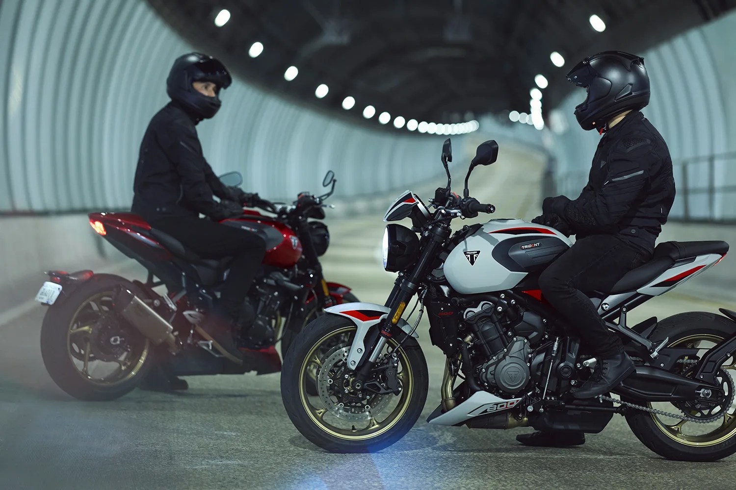 Two motorcyclists wearing black helmets and jackets sitting on a white and a red Triumph Trident 660 motorcycle in a tunnel.