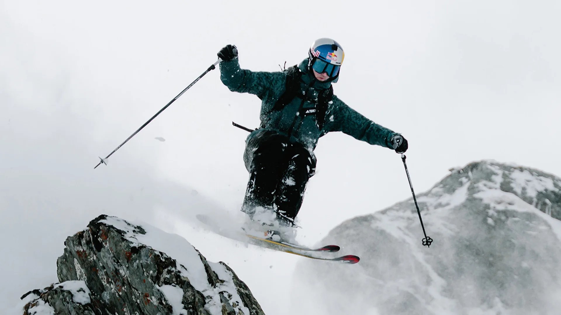 Skier in dark green jacket and black pants jumping off a snowy rocky ledge with ski poles in hand.