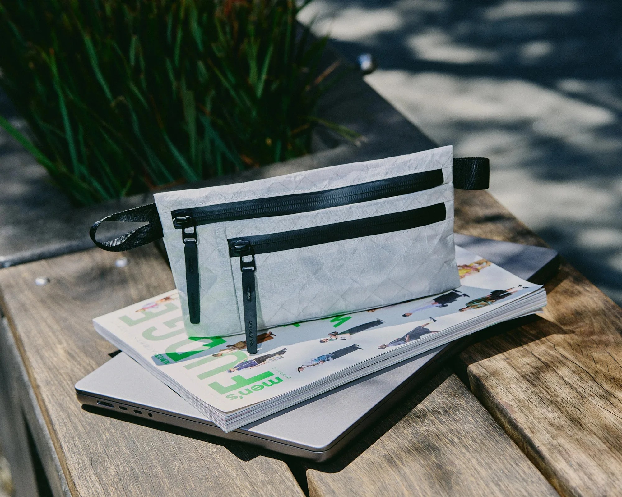Light gray rectangular pouch with two black zippers and black pull tabs, placed on a magazine and closed laptop on a wooden bench.