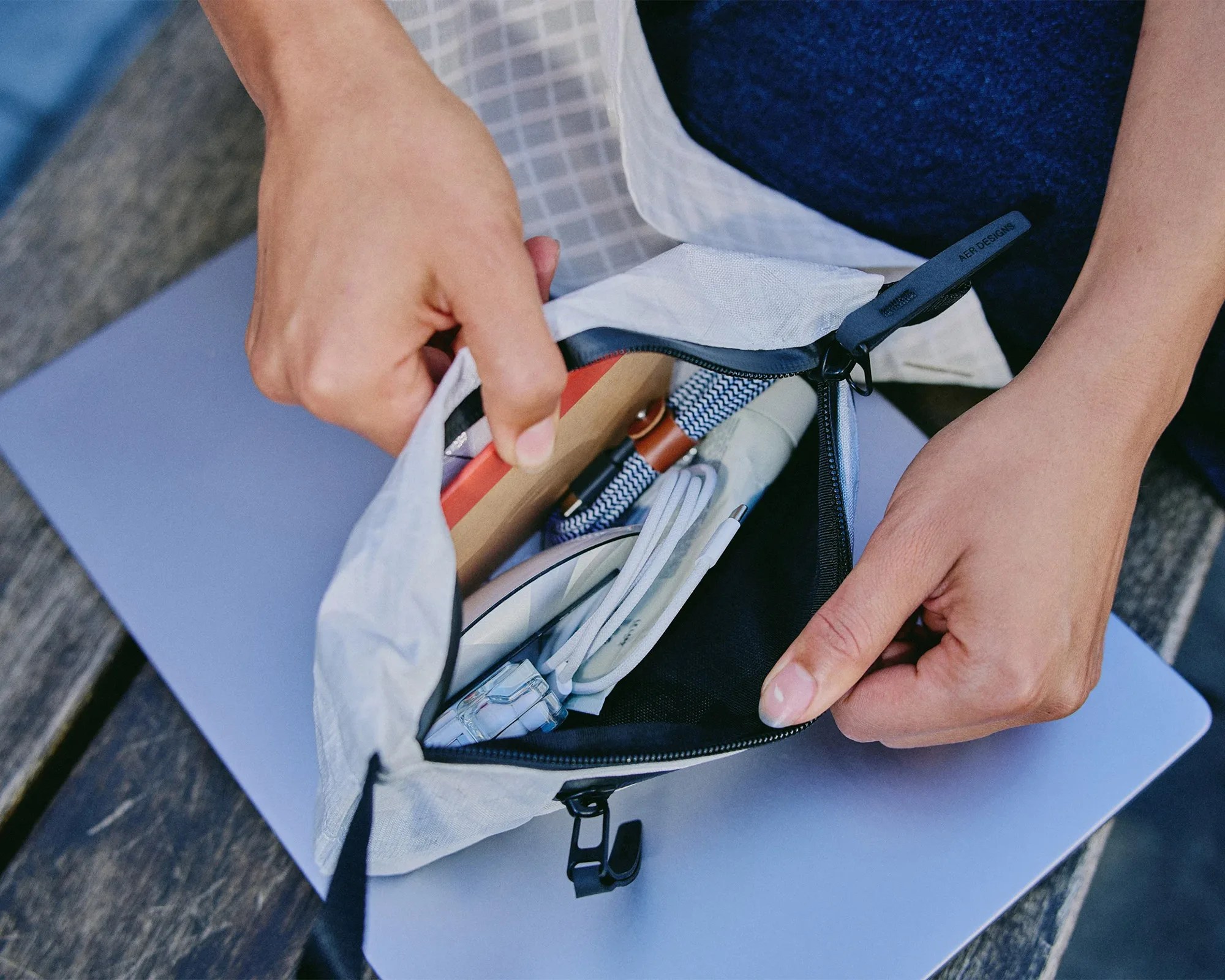 Hands holding open a small white zippered pouch containing a notebook, cable, and other small items on a laptop.