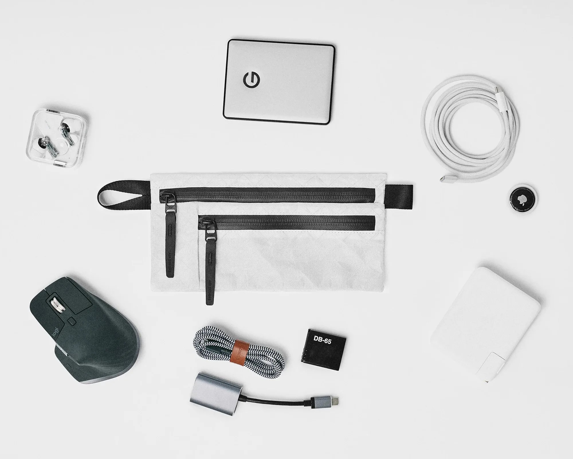 White fabric pouch with two black zippers surrounded by various tech accessories on a white surface.