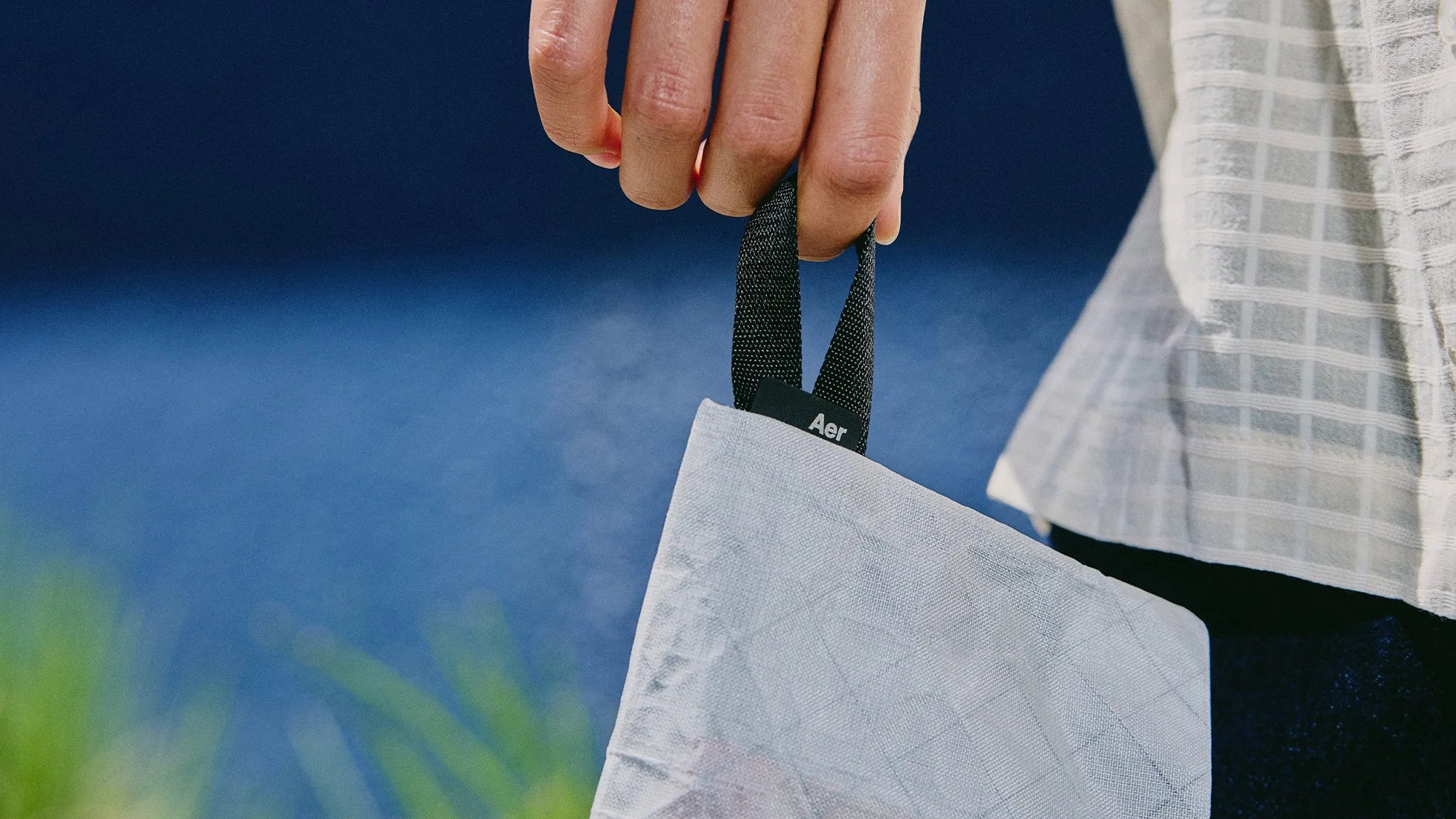 Hand holding a light gray textured tote bag with black handles and an "Aer" label against a blue background.