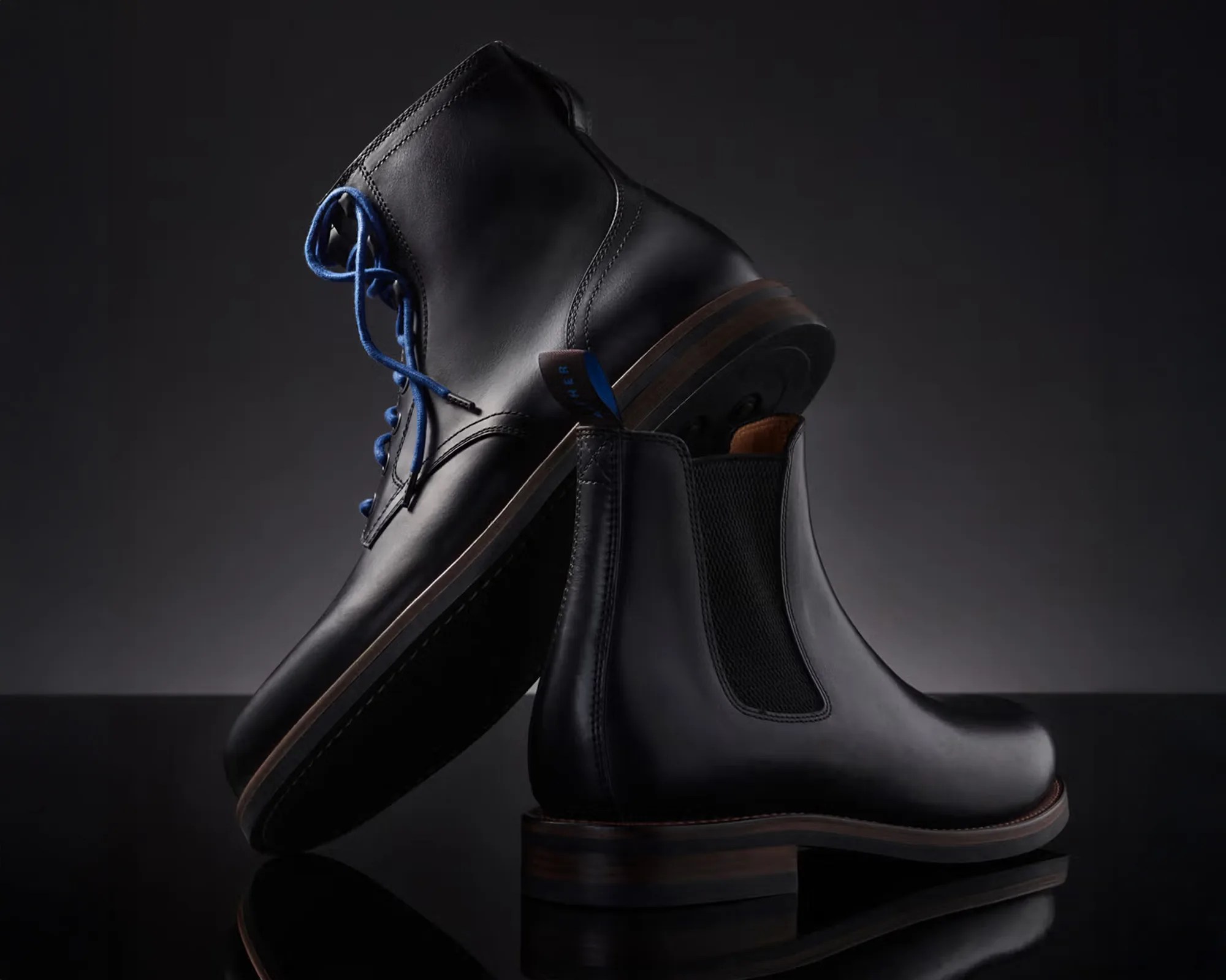 Pair of black leather boots, one with blue laces and the other a slip-on style with elastic side panels, on a reflective black surface.