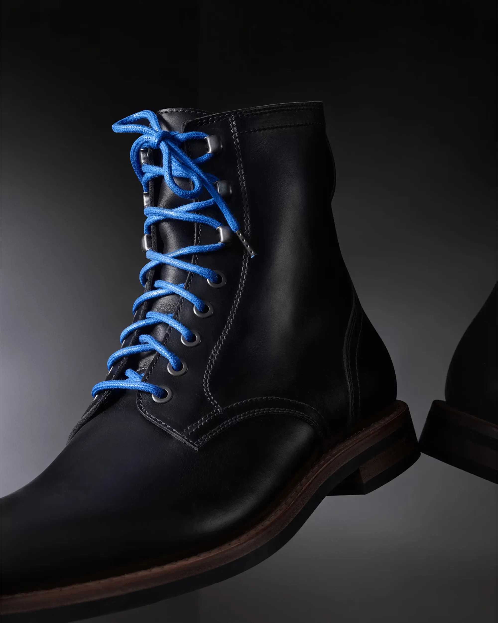 Black leather ankle boot with bright blue laces and brown sole on a dark background.
