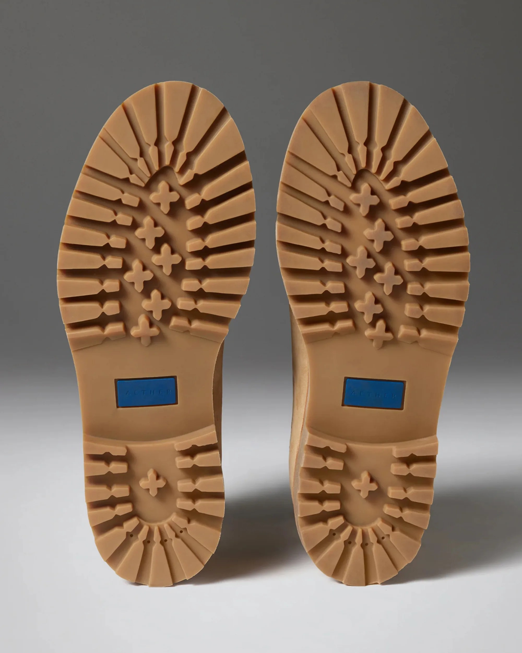 Pair of tan rubber boot soles with deep treads and blue rectangular logo inserts.