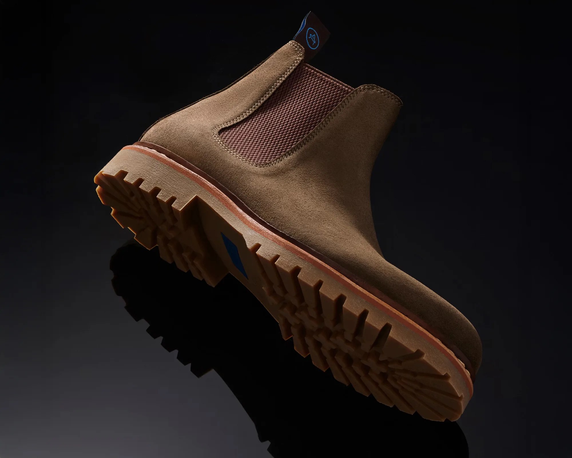 Brown suede Chelsea boot with lug sole and elastic side panel on a black reflective surface.