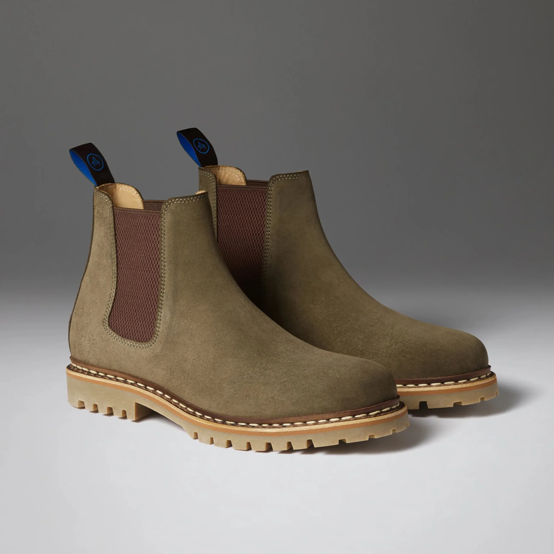Pair of olive green suede Chelsea boots with brown elastic side panels and beige lug soles on a gray surface.