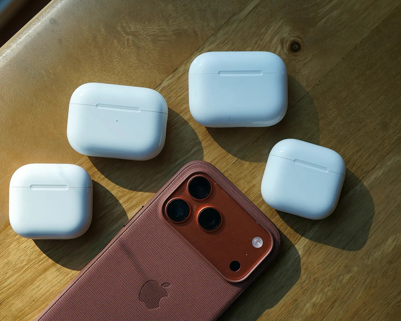 Copper-colored iPhone with triple camera lenses and four white wireless earbud charging cases on a wooden surface.