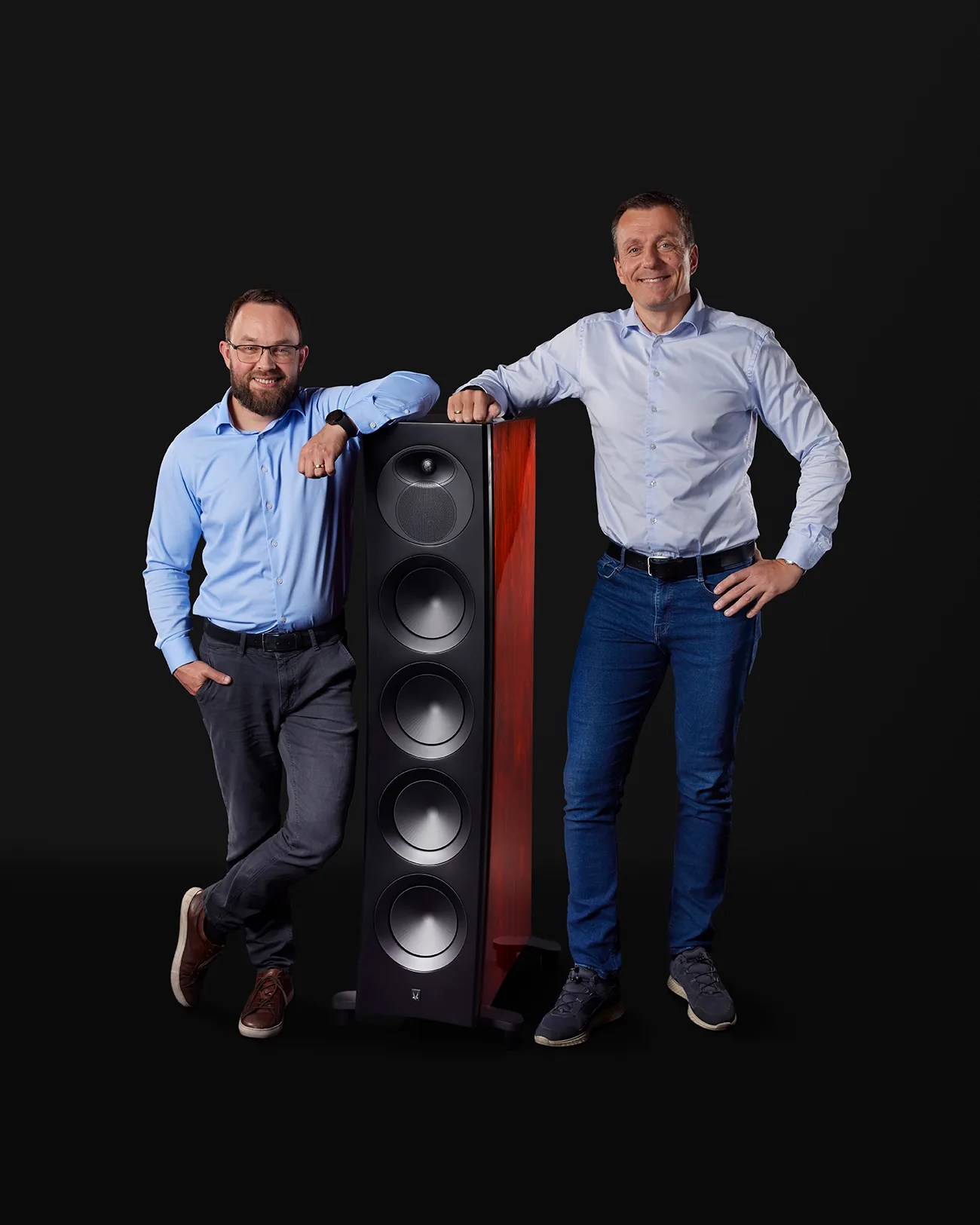 Two men in light blue shirts and dark pants standing on either side of a tall black and wood-finished floor-standing speaker against a black background.