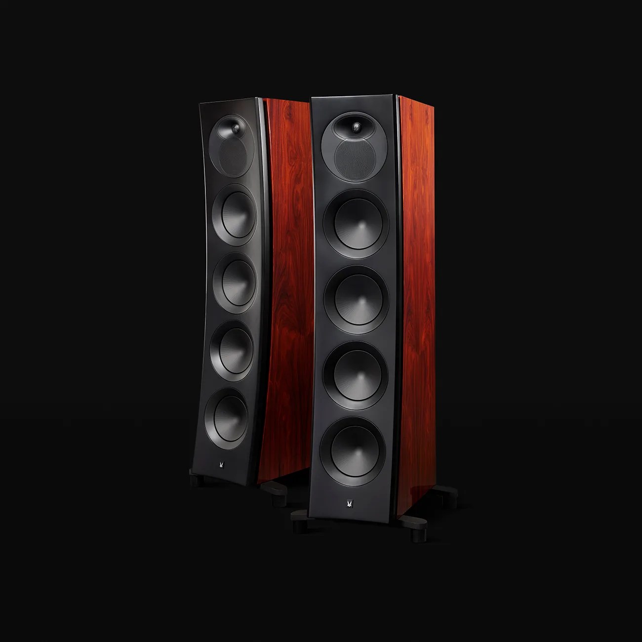 Pair of tall floor-standing speakers with black front panels and reddish wood side panels on black stands.