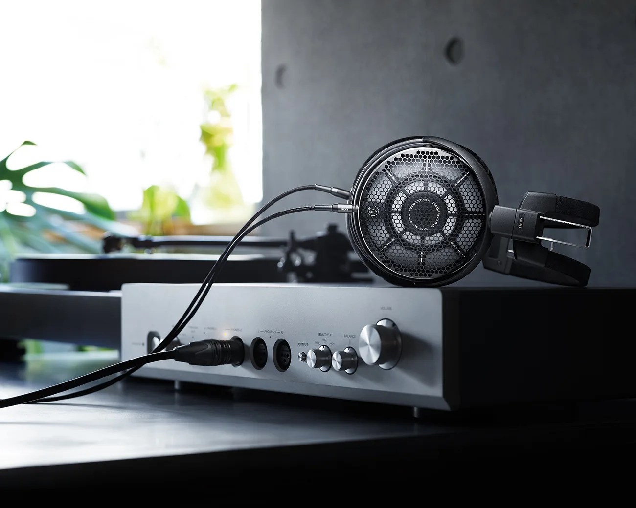 Black open-back headphones connected to a silver audio amplifier on a dark surface.