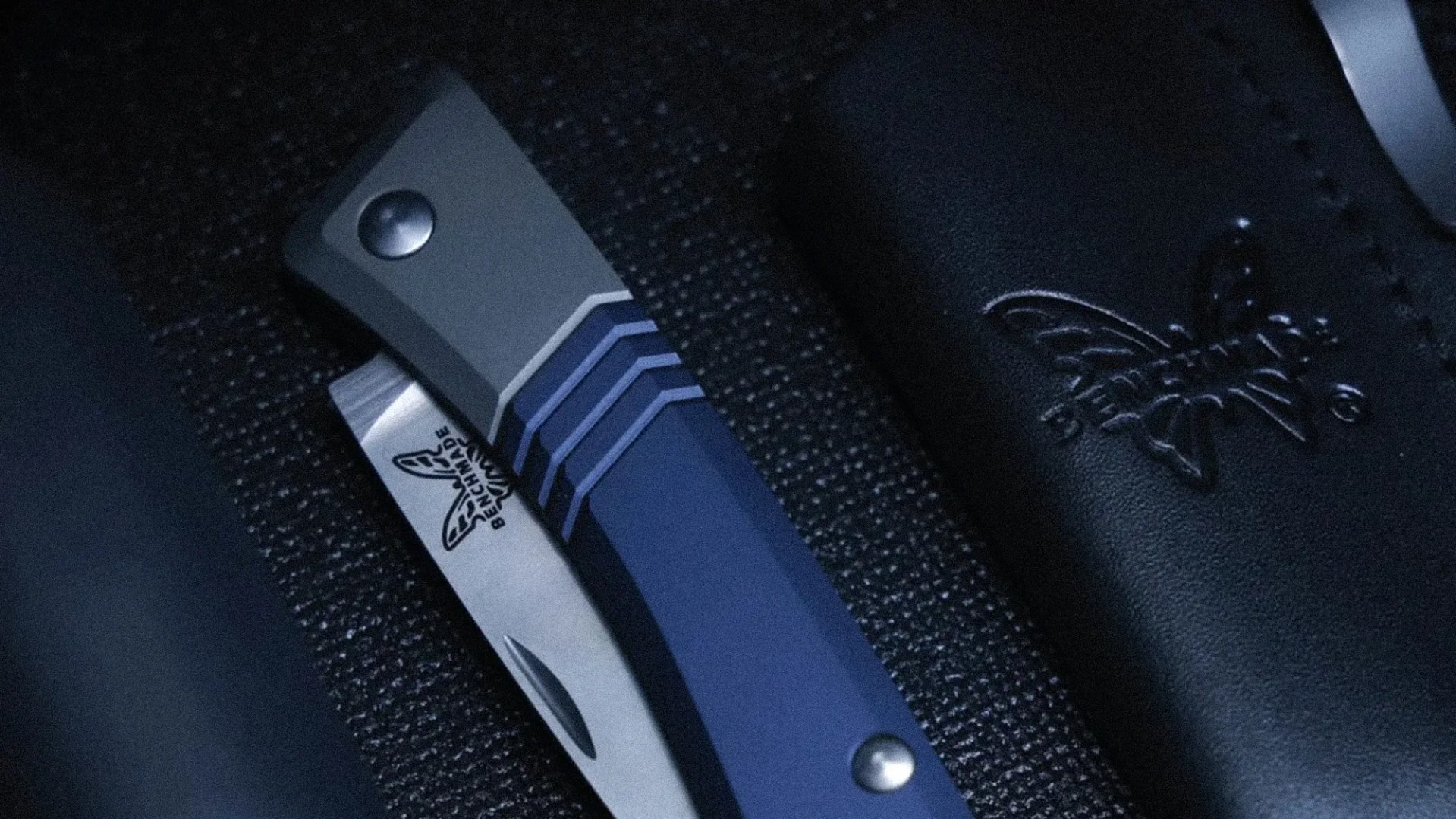 Folding knife with blue and gray handle and partially visible blade engraved with Benchmade logo on textured black surface.