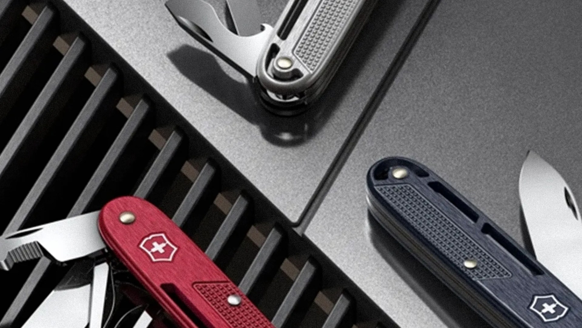 Three Swiss Army knives in red, silver, and blue with partially opened blades on a textured gray surface.