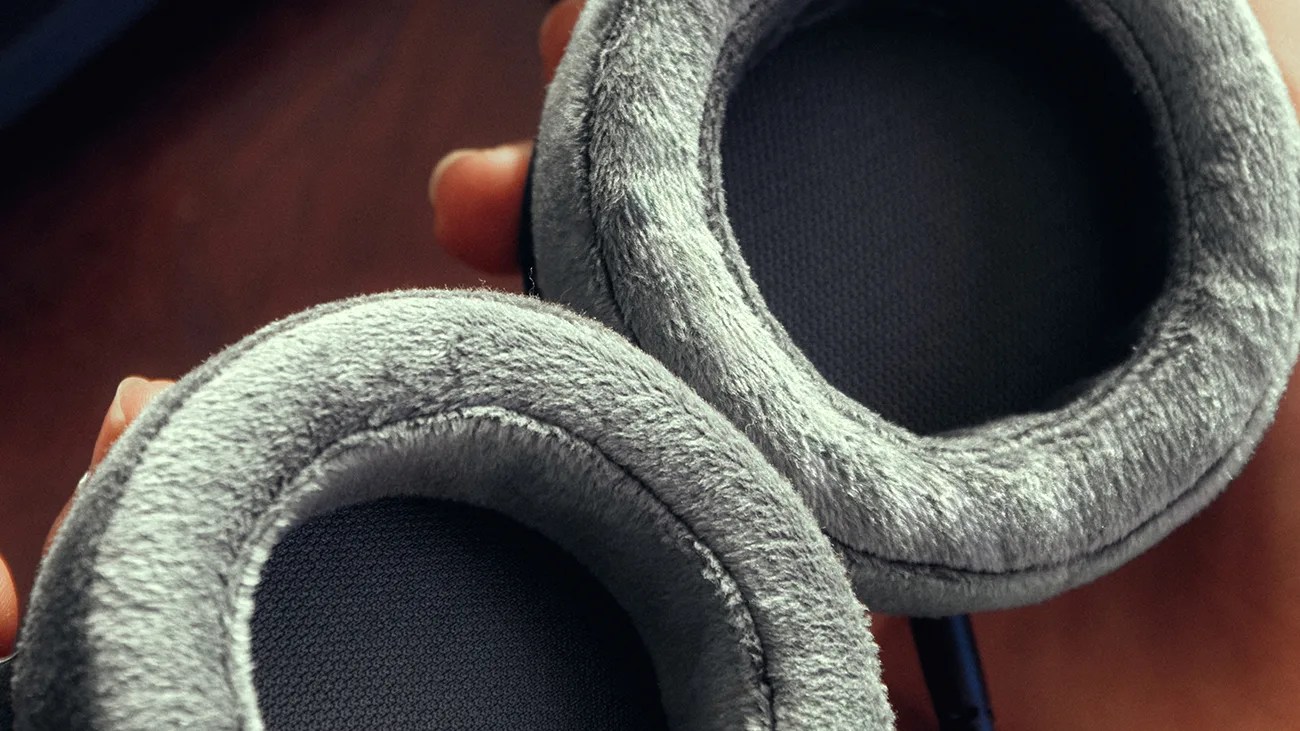 Close-up of gray plush headphone ear cushions held in a hand against a blurred background.
