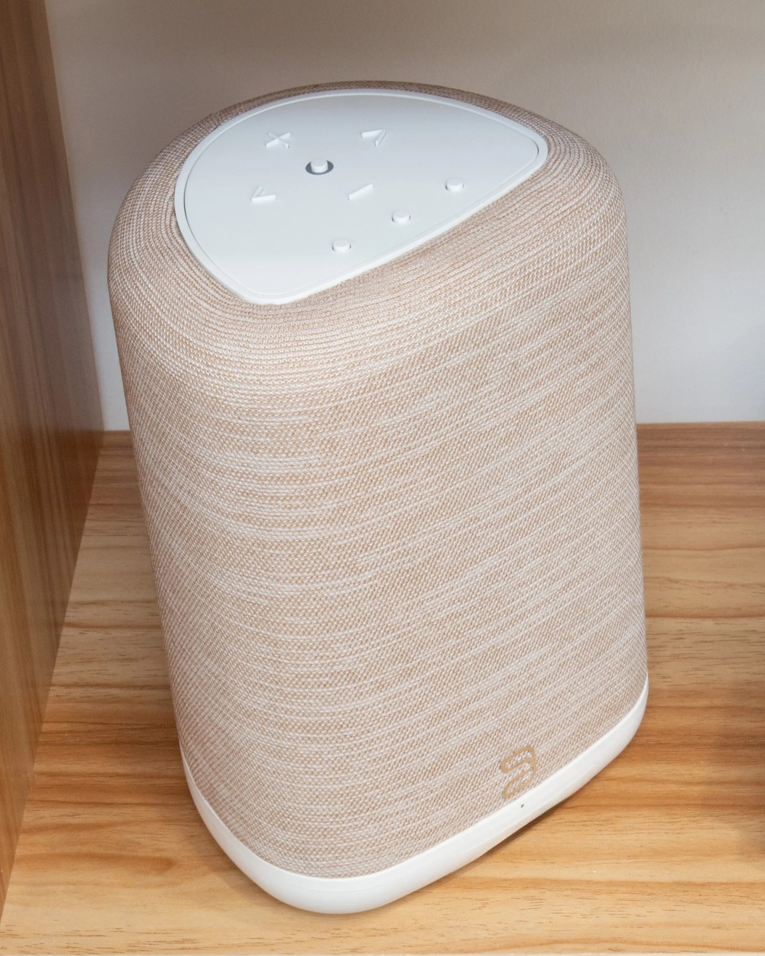 Beige fabric-covered smart speaker with white top control panel on a wooden surface.