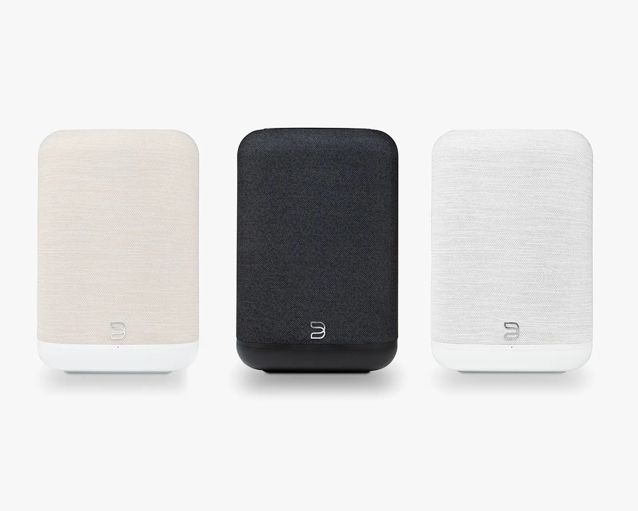 Three rectangular smart speakers with rounded edges in beige, black, and white fabric covers, each with a small logo near the bottom.