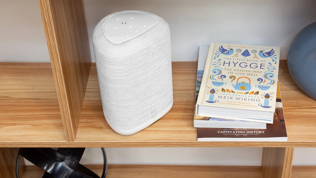 White fabric-covered smart speaker with touch controls on top placed on a wooden shelf next to a stack of books.