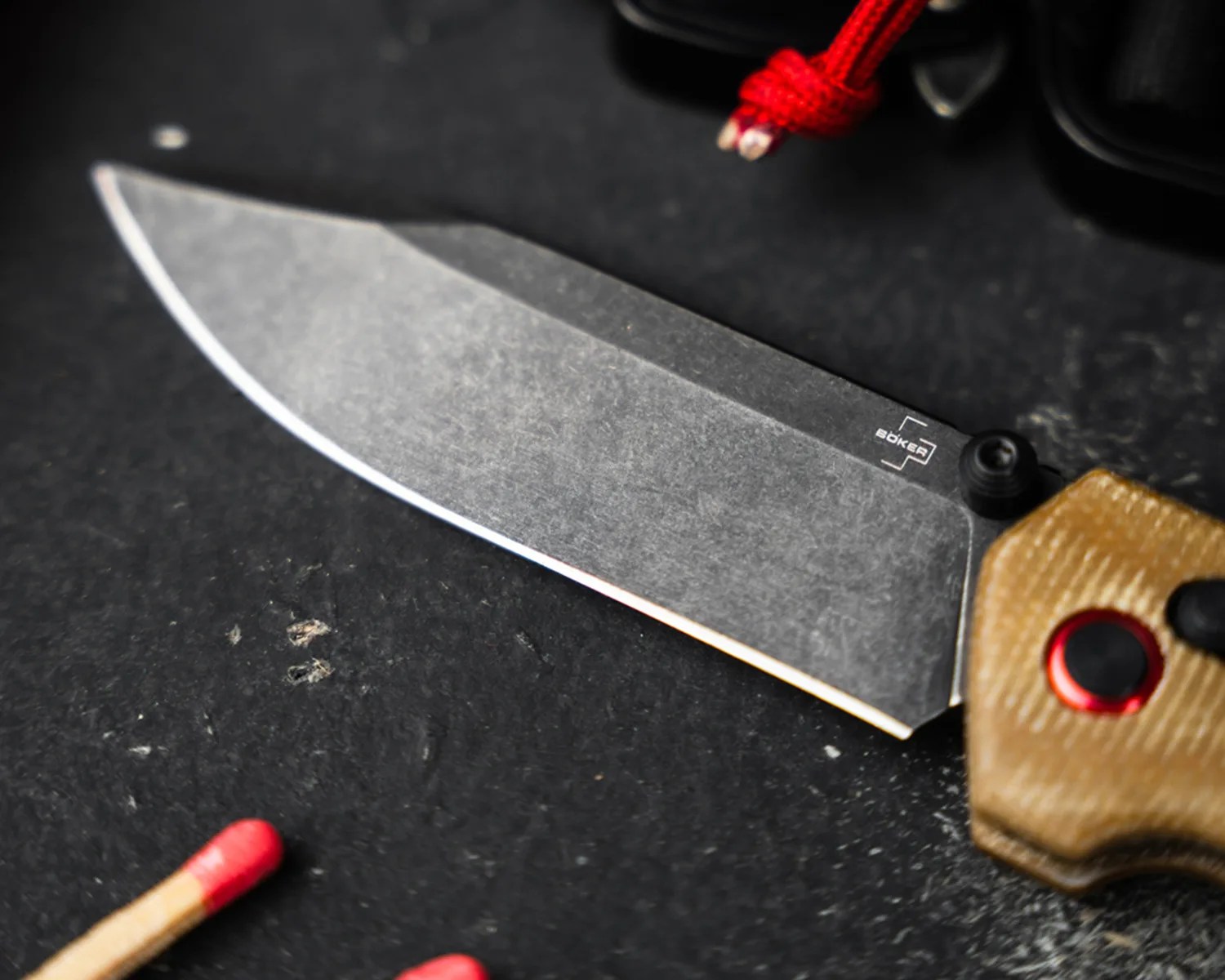Stonewashed blade of a Boker folding knife with tan textured handle and red-accented button on a black surface.