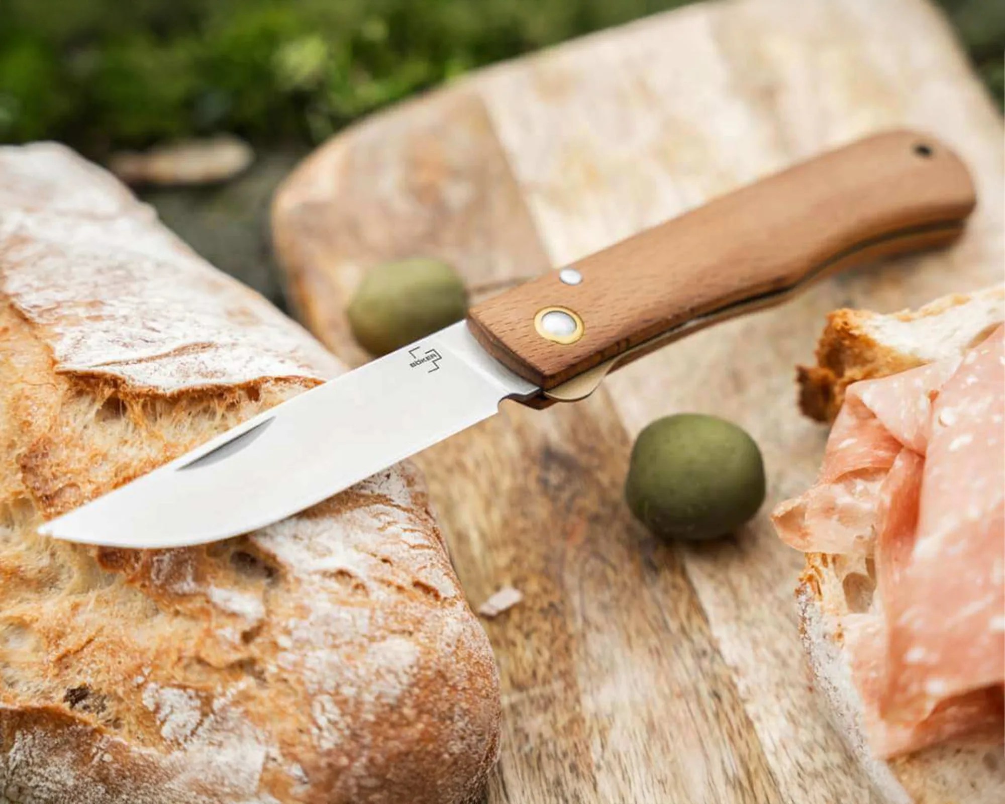 Folding knife with a wooden handle and stainless steel blade on a wooden cutting board with bread, olives, and sliced meat.