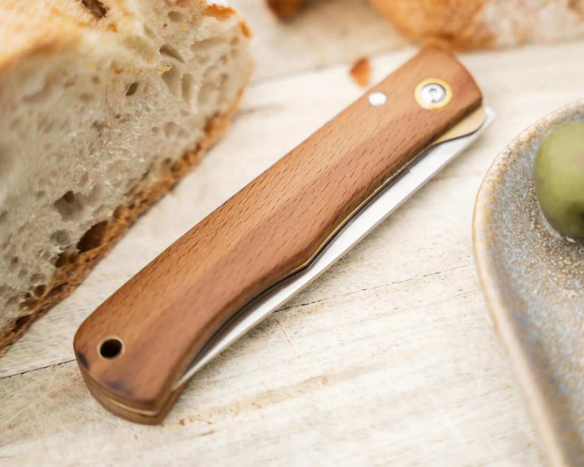 Closed folding knife with a wooden handle on a light wooden surface next to bread and a plate.