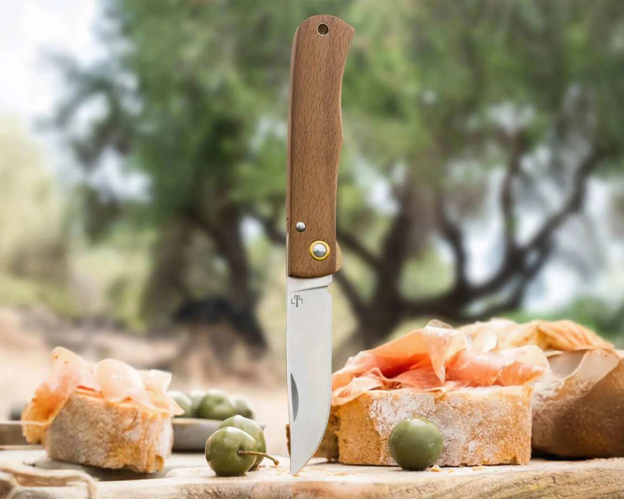 Folding knife with wooden handle stabbed into wooden board with bread, prosciutto, and green olives.