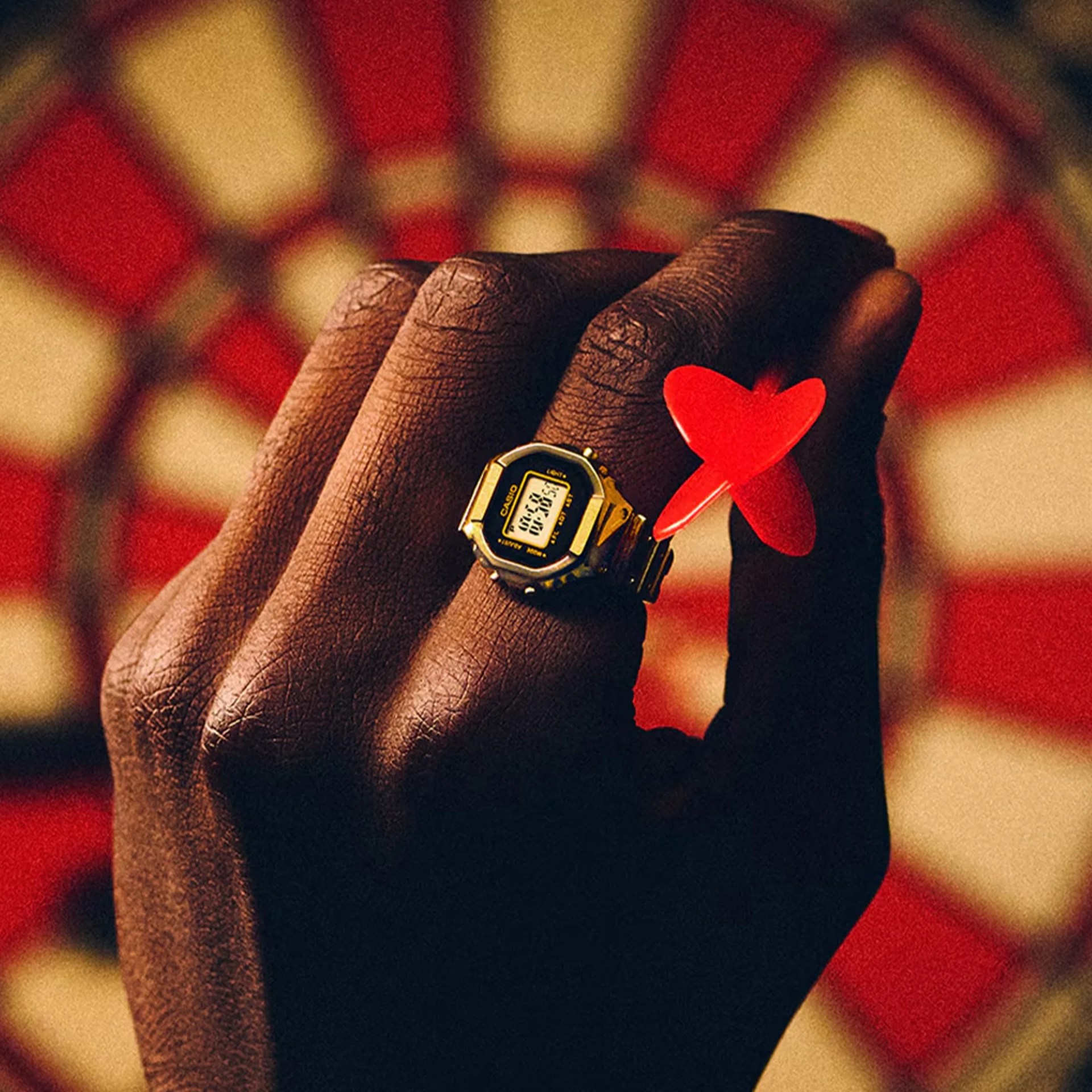 Hand with dark skin wearing a gold Casio digital watch and holding a red dart in front of a dartboard.