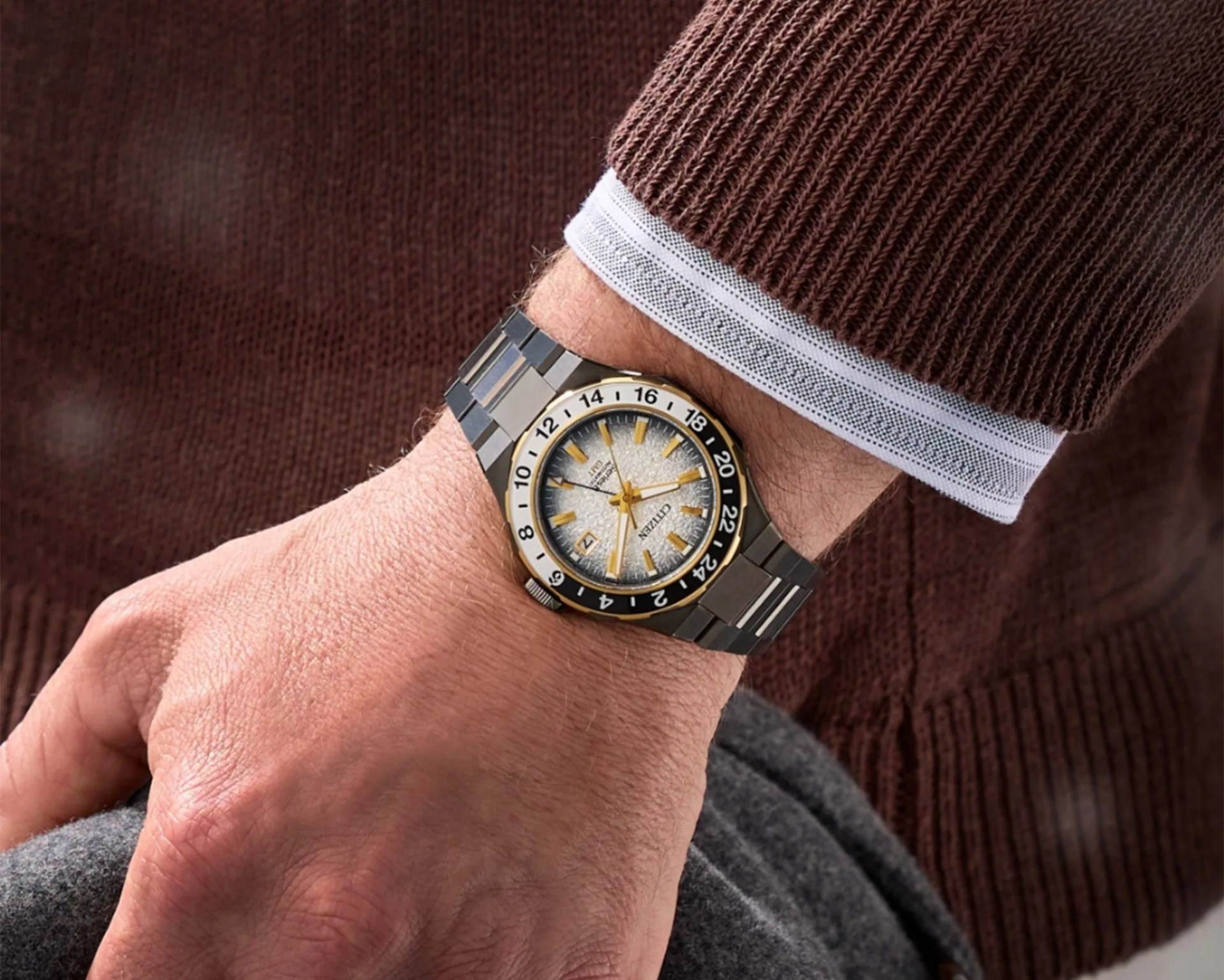 Silver and gold Citizen wristwatch with black and white 24-hour bezel on a wrist with a brown sweater.