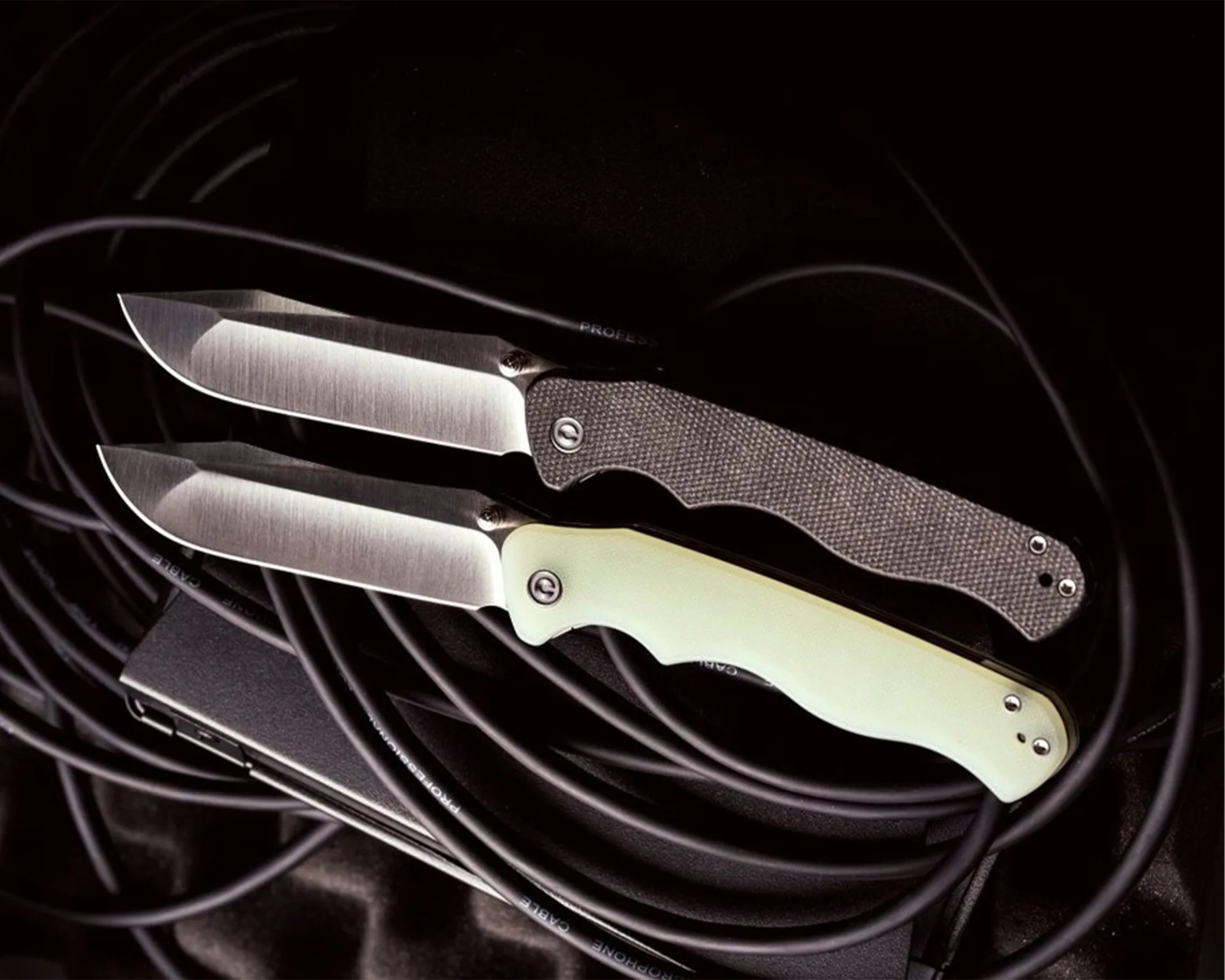 Two folding knives with satin-finished blades, one with a black textured handle and the other with a smooth pale green handle, on black cables.
