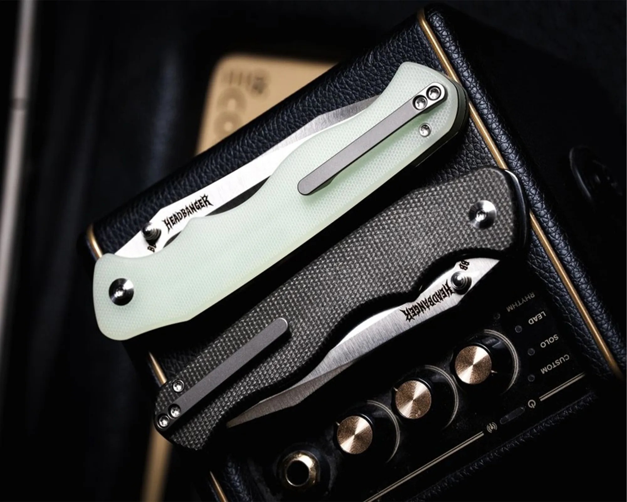 Two closed folding knives with "HEADBANGER" engraved on the blades, one with a light green handle and the other with a textured gray handle, resting on a black amplifier.