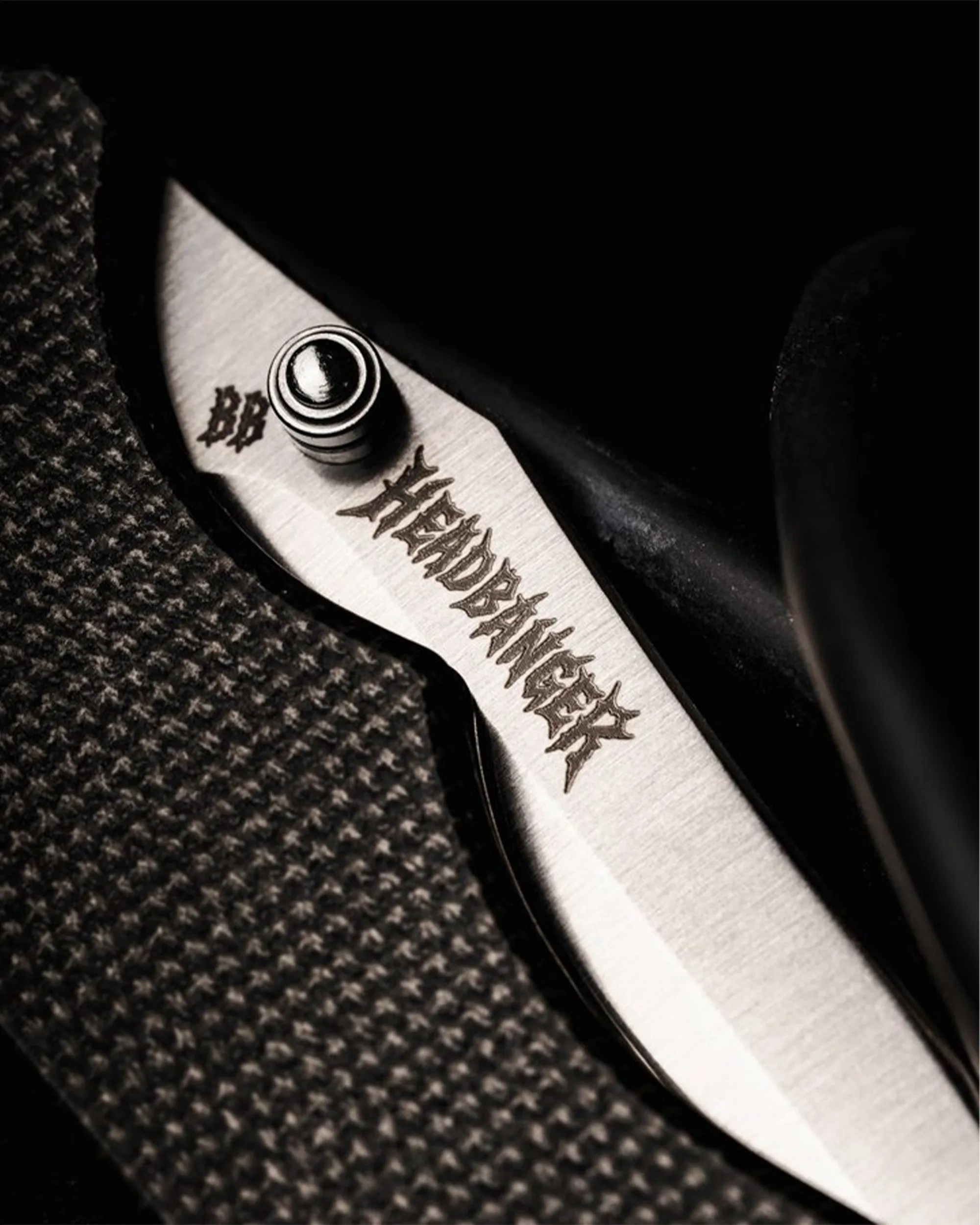 Close-up of a folding knife blade with "HEADBANGER" engraved near the pivot screw and a textured black handle.