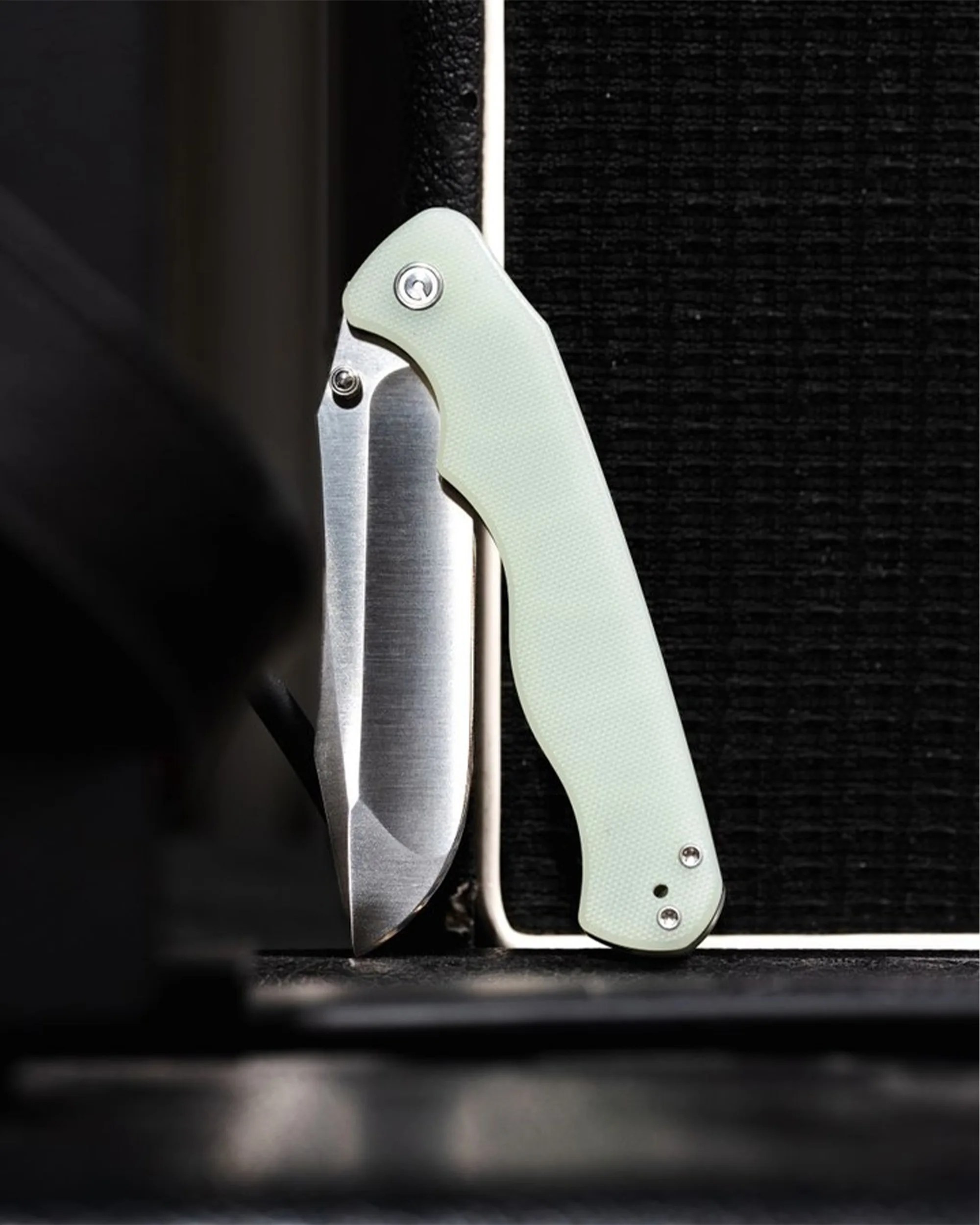 Folding knife with a partially open silver blade and a pale green textured handle standing upright on a dark surface.