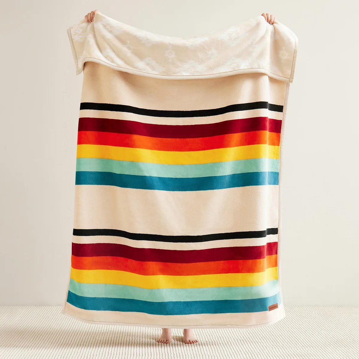 Cream blanket with horizontal stripes in black, burgundy, orange, yellow, light blue, and teal held up by a person.