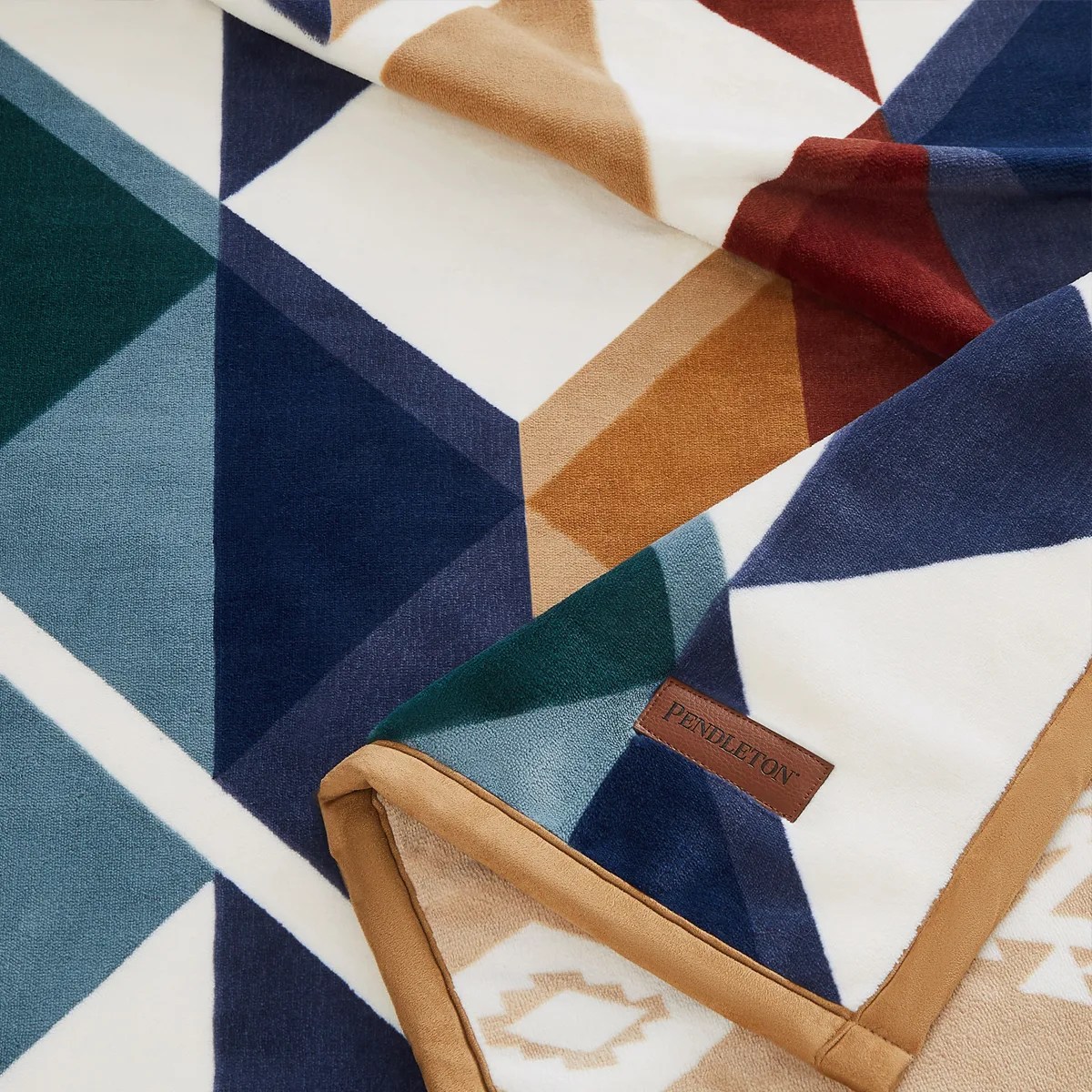 Geometric patterned blanket in navy, teal, rust, tan, and white with a tan border and Pendleton leather tag.