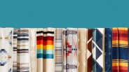 Row of folded blankets with various geometric and striped patterns in colors like beige, red, blue, yellow, and orange against a teal background.