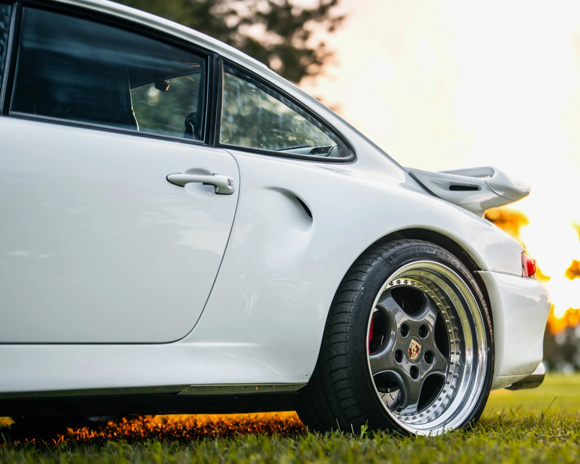 White Porsche sports car rear side with polished multi-piece alloy wheel on grass at sunset.