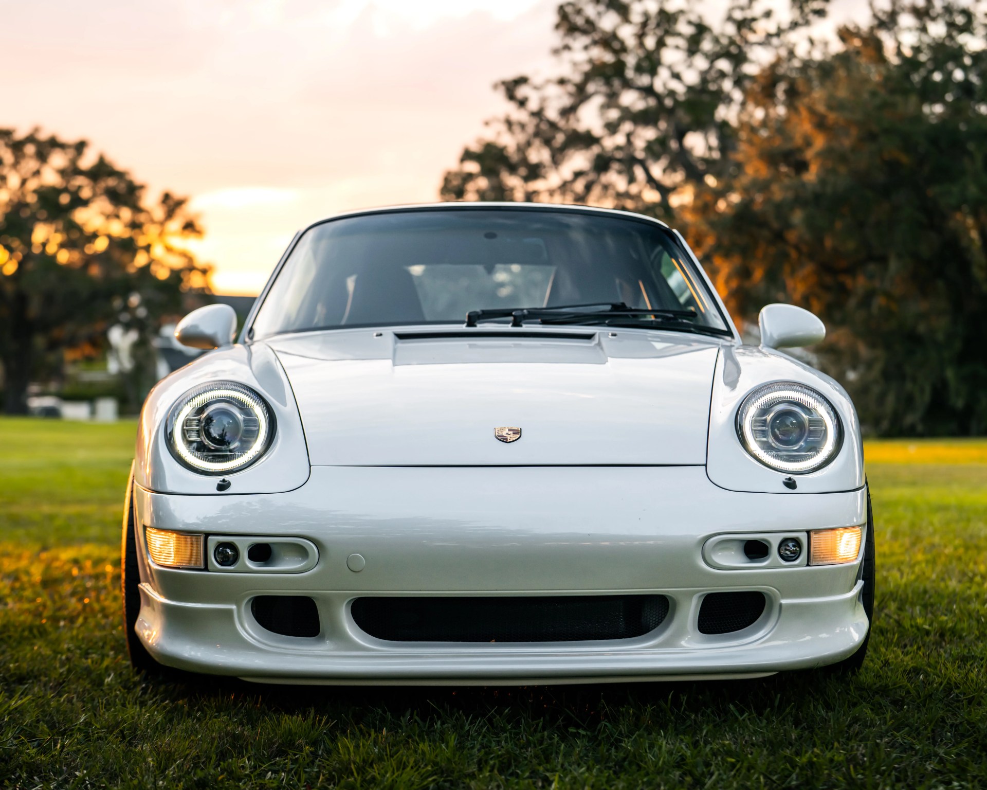 Front view of a white Porsche sports car with illuminated round headlights parked on grass at sunset.