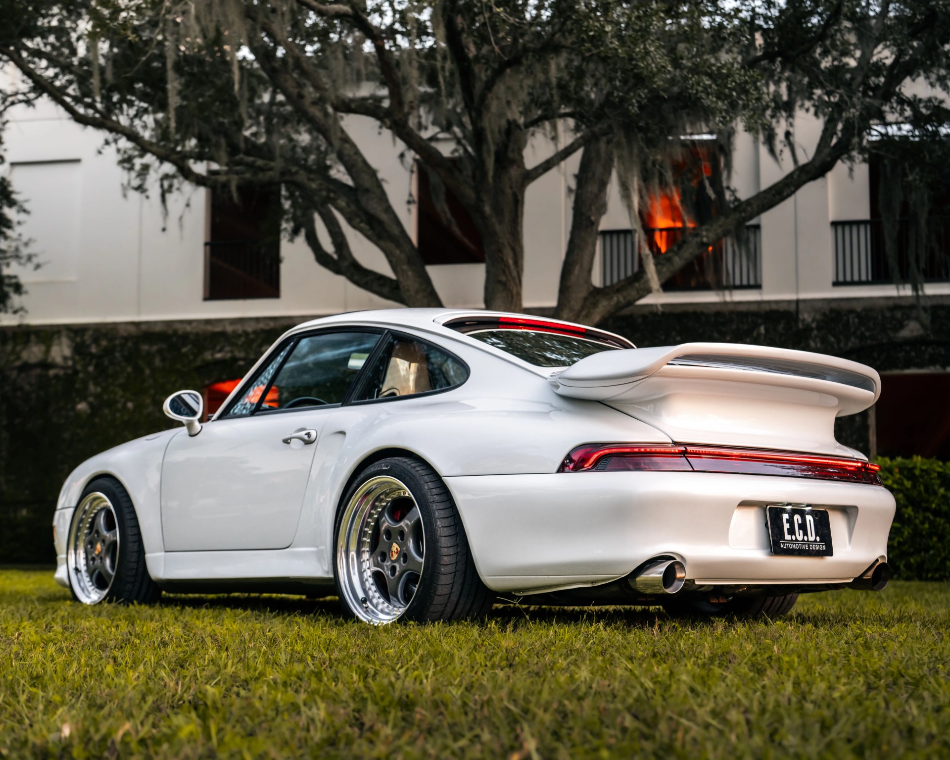 White Porsche 911 with wide rear spoiler and polished deep-dish wheels parked on grass.