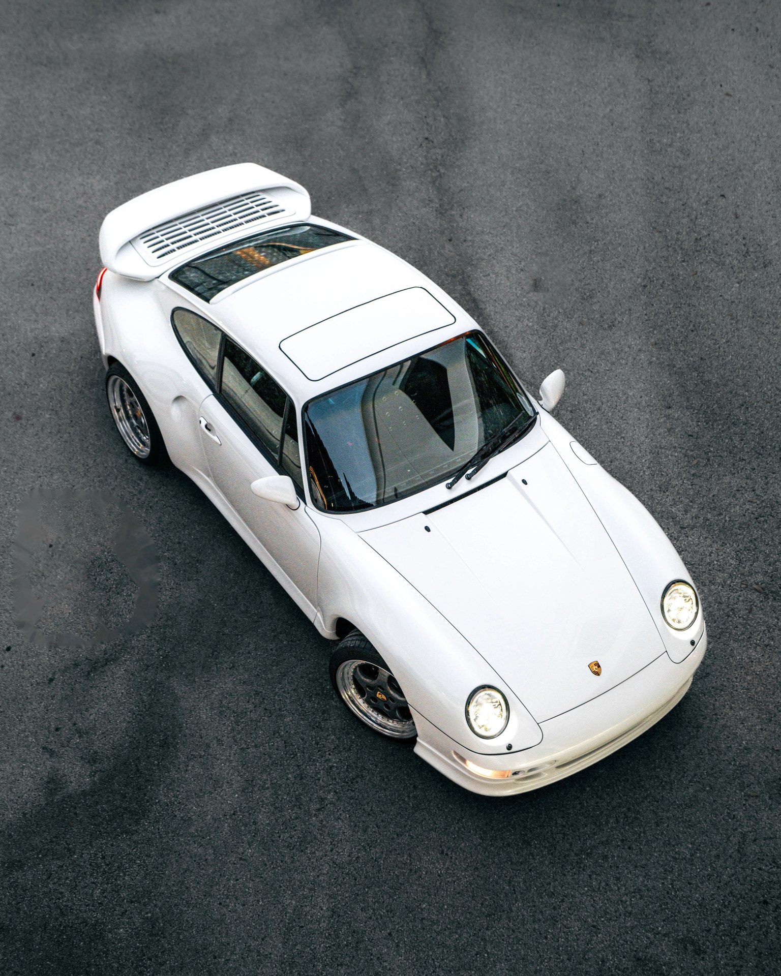 White Porsche 911 sports car with rear spoiler parked on gray asphalt, viewed from above.