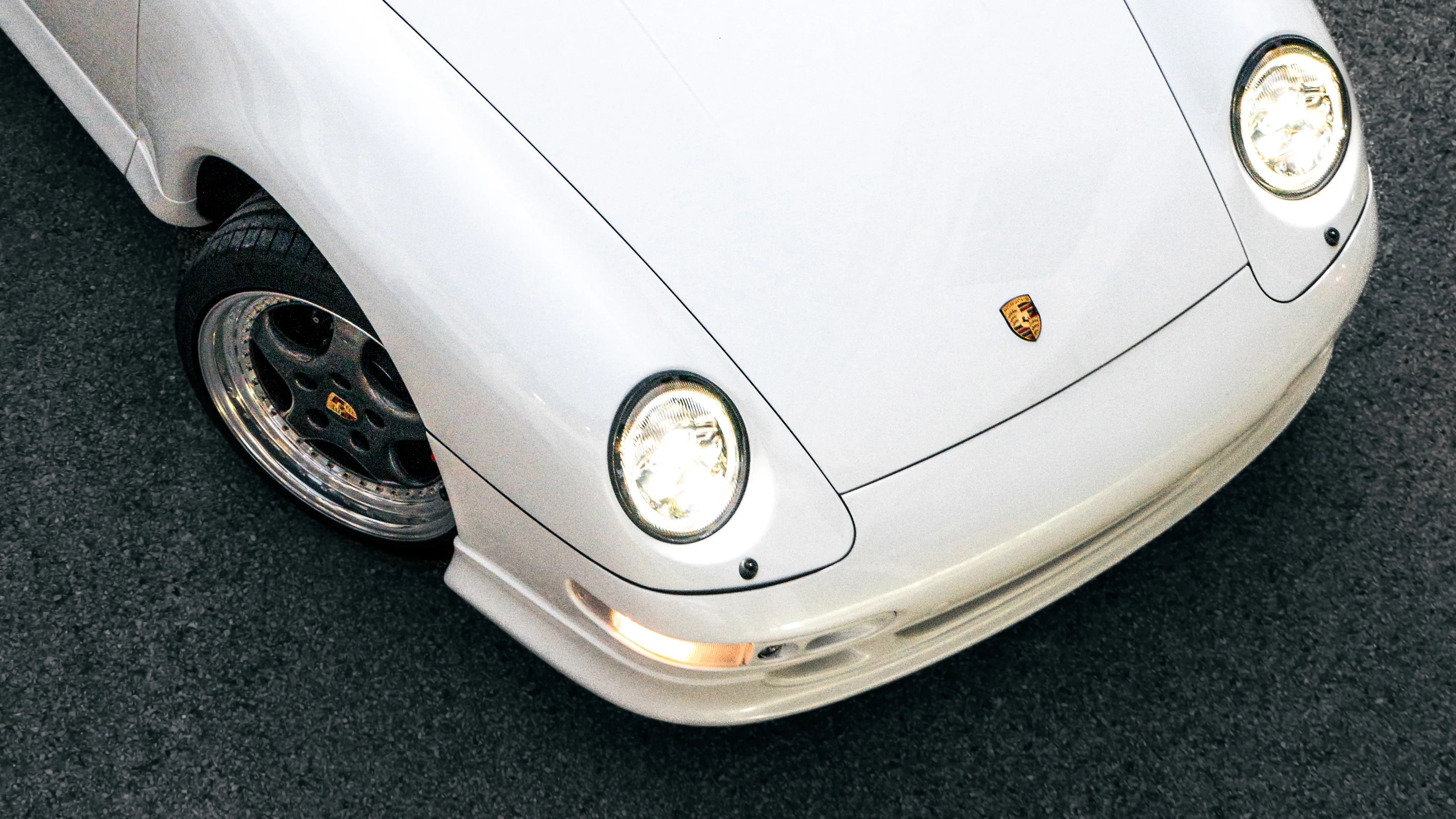 Front corner of a white Porsche sports car with headlights on and black rims on asphalt.