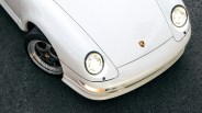 Front corner of a white Porsche sports car with headlights on and black rims on asphalt.