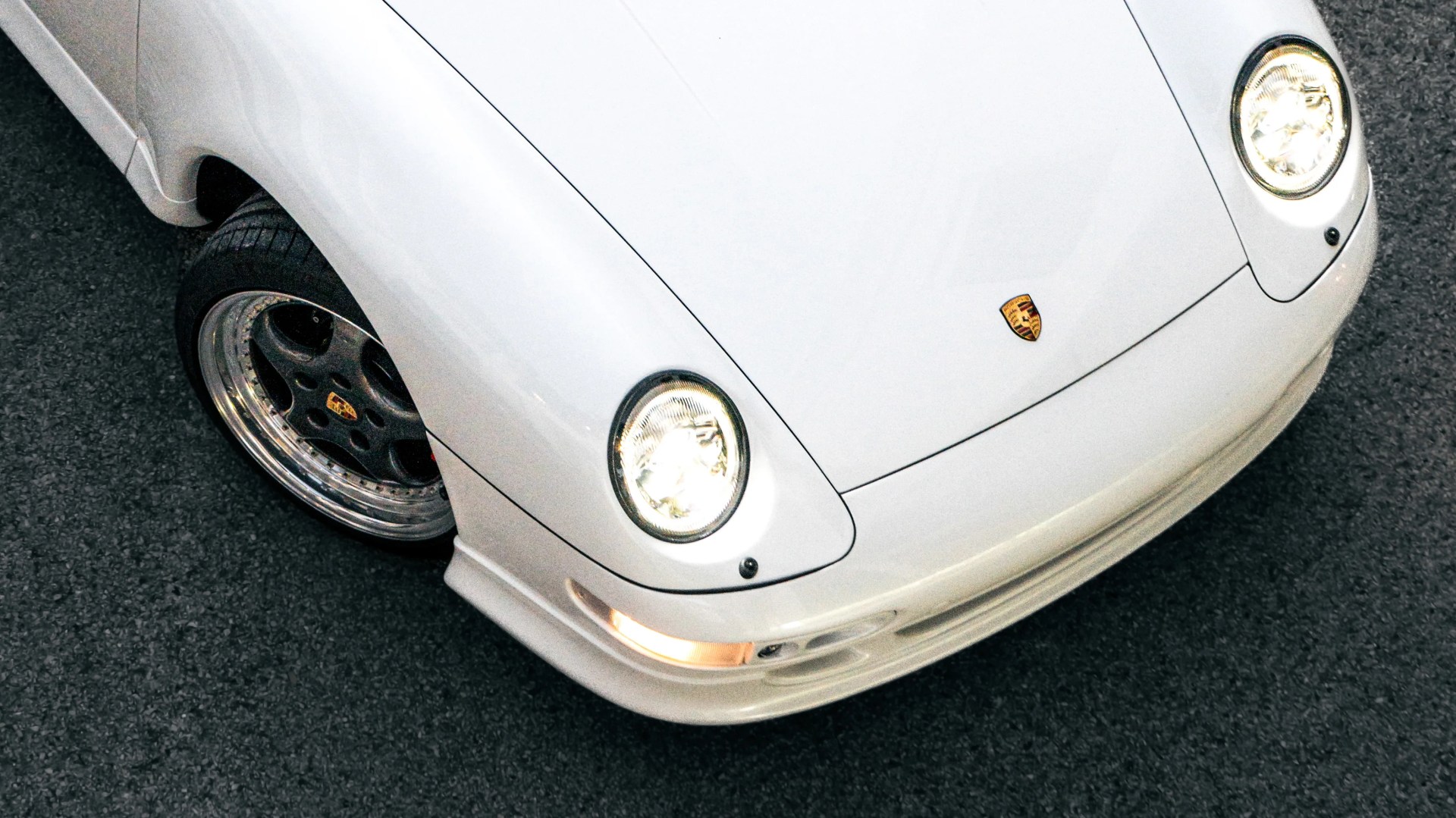 Front corner of a white Porsche sports car with headlights on and black rims on asphalt.