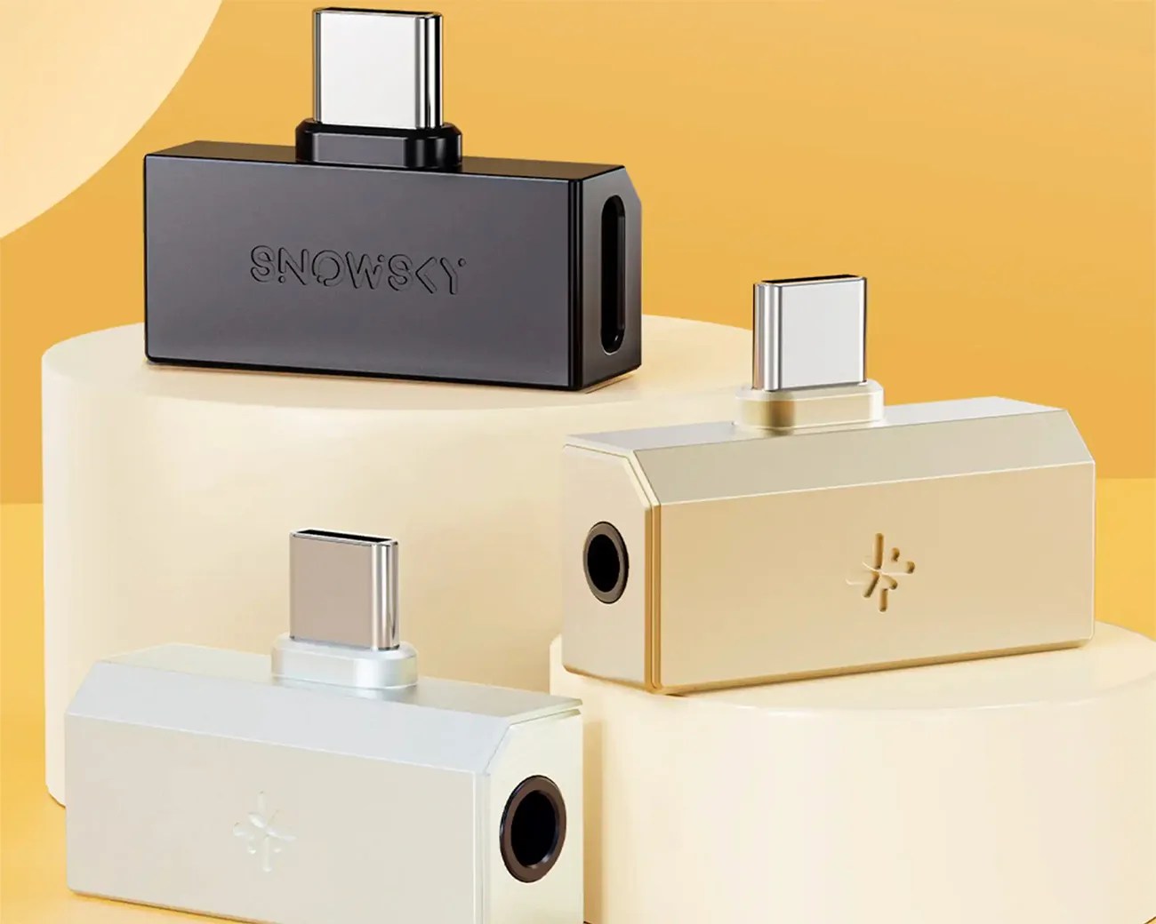 Three rectangular USB-C to 3.5mm headphone jack adapters in black, gold, and silver on cream-colored cylindrical stands.