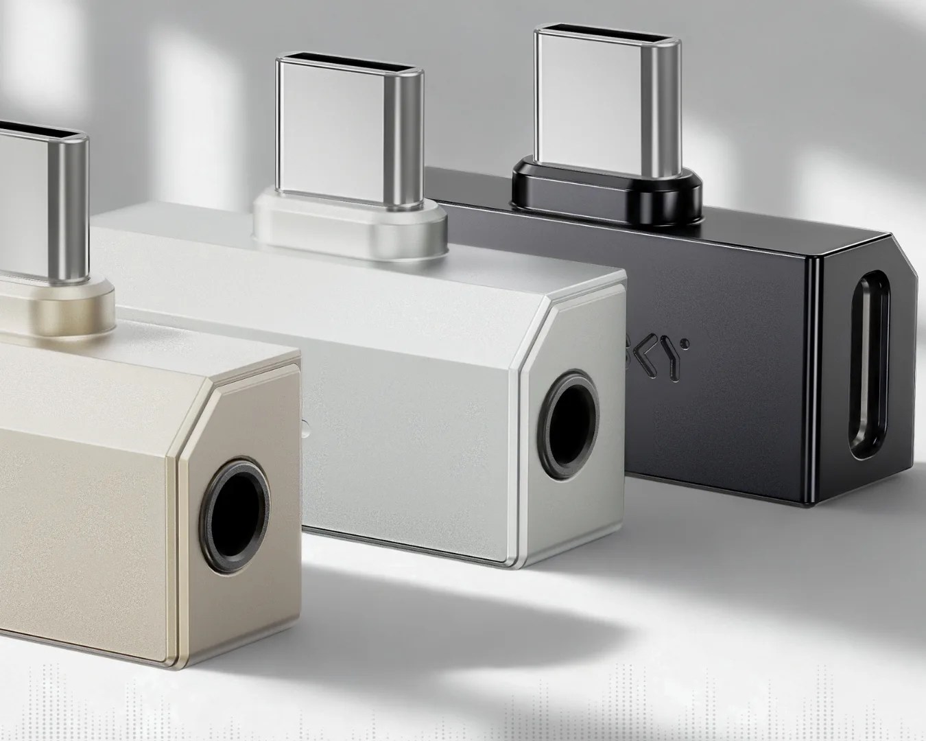 Three rectangular USB-C to 3.5mm headphone jack adapters in beige, white, and black on a gray surface.