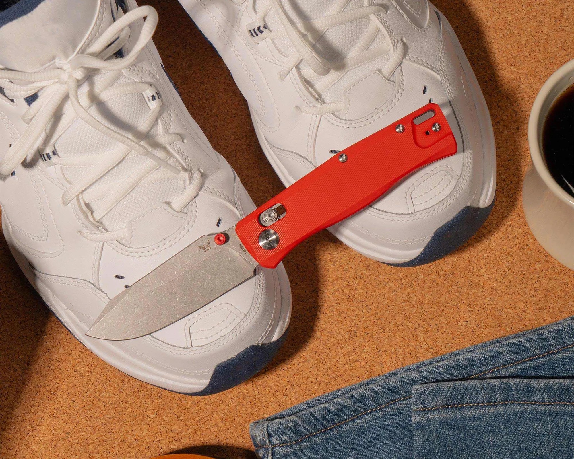 Stonewashed blade folding knife with bright orange handle resting on white sneakers on a cork surface.