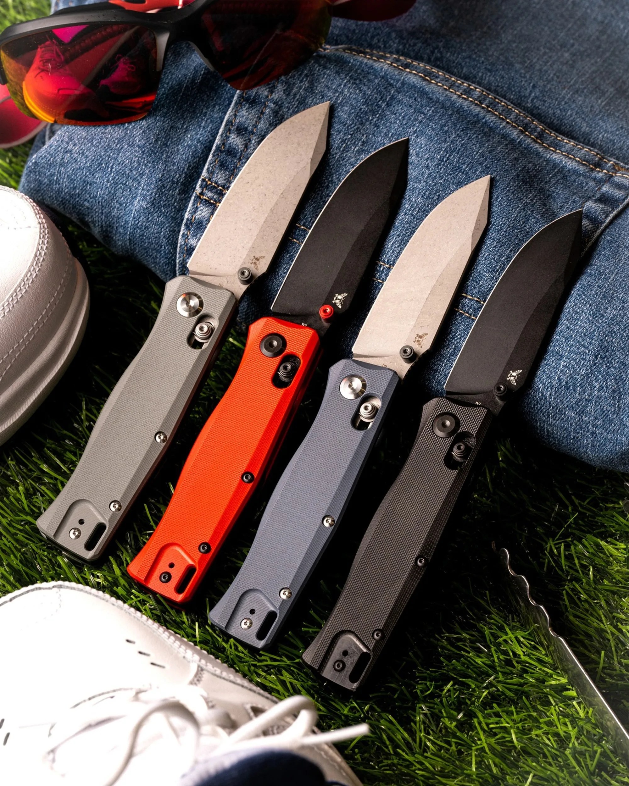 Four folding knives with textured handles in gray, orange, blue, and black, laid open on grass next to white sneakers, sunglasses, and denim.