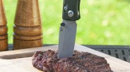 Gray folding knife blade stabbed into grilled steak on a wooden cutting board outdoors.